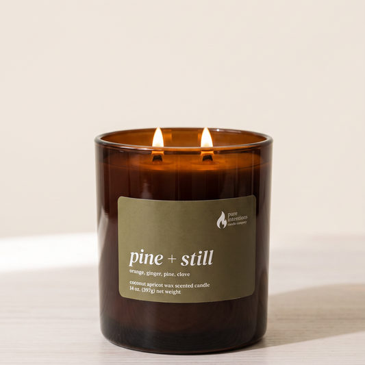 An amber glass jar candle with two lit wicks and a muted olive-green label reading 'pine + still' on a light beige background.
