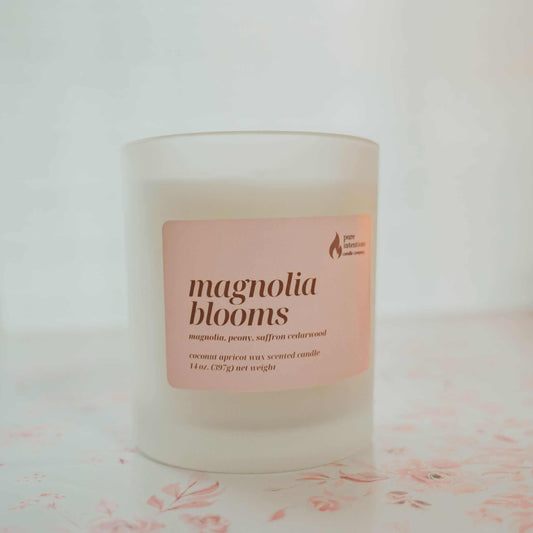 Candle labeled ' magnolia blooms' on a white background with pink flower petals.
