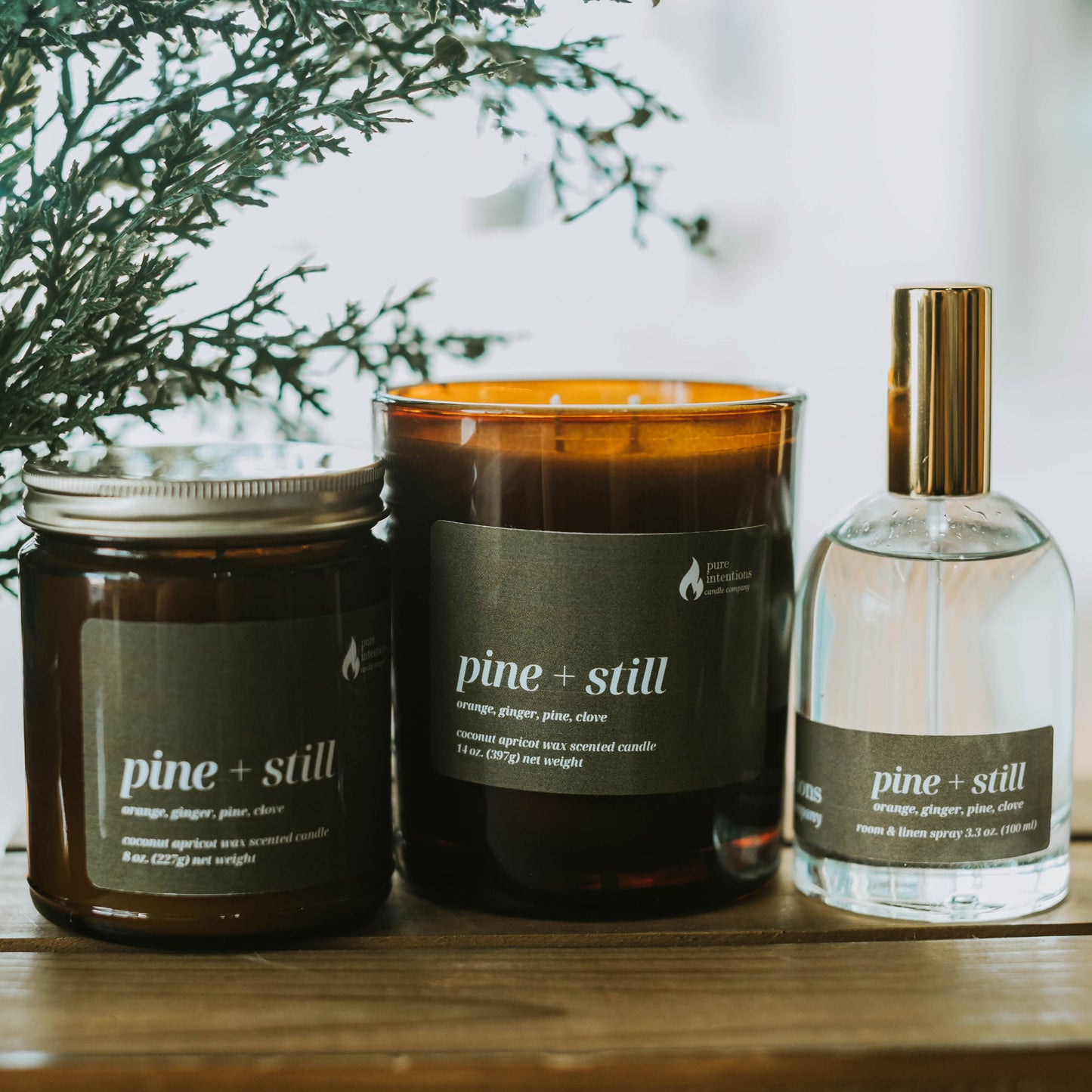 Three 'pine + still' products including a candle, a jar, and a spray bottle on a wooden surface with a blurred natural background.