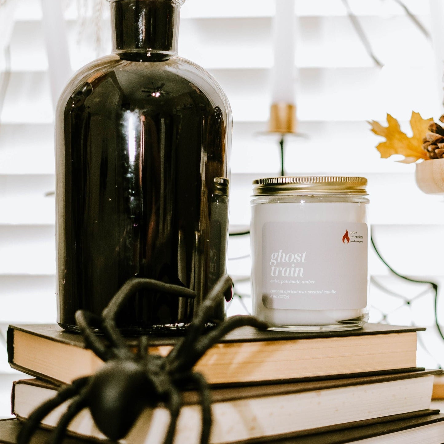 A black jar labeled 'Ghost Train' by Pure Intention Candle Co. is displayed on a table with a decorative spider, books, and a pine cone in the background.