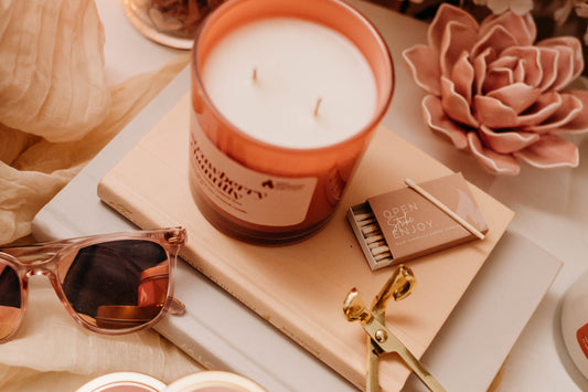 A cozy self-care setup featuring a Strawberry Chantilly candle from Pure Intentions Candle Company, placed on a book with a matchbox, gold wick trimmer, and soft decor accents including pink flowers and sunglasses.