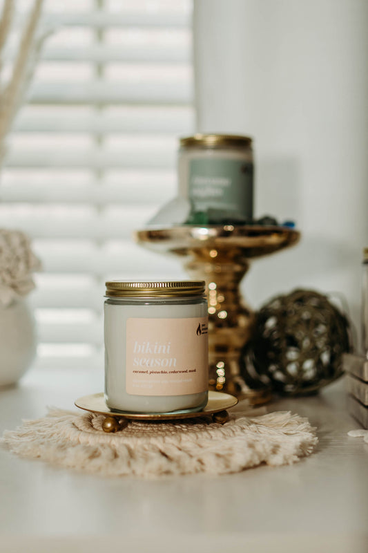 The image displays two candles from the Pure Intentions Candle Company's "Bikini Season" collection. The front candle is labeled "Bikini Season" with scents of caramel, pistachio, cedarwood, and musk. It sits on a decorative, round woven mat atop a gold-colored stand. In the background, another candle labeled "Havana Nights" is slightly blurred, also placed on a decorative stand. The setting is light and airy, with soft focus elements adding a cozy, bohemian ambiance.