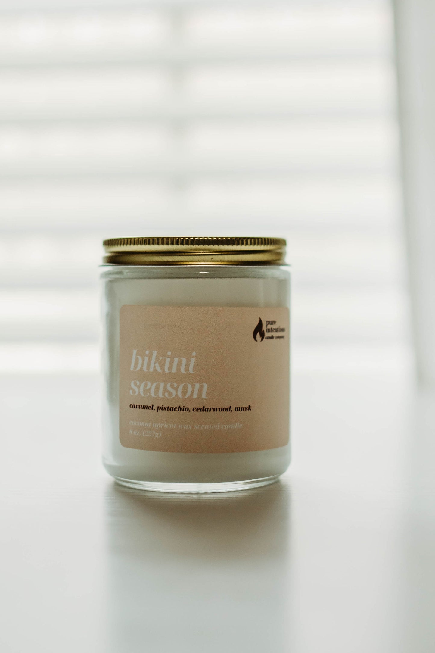 The image features a single candle from Pure Intentions Candle Company's "Bikini Season" collection. The candle is housed in a clear glass jar with a gold-colored lid. The label is light pink with white text, stating the scent as "Bikini Season" and listing the fragrance notes of caramel, pistachio, cedarwood, and musk. The background is softly lit with blurred white shades, creating a calm and serene atmosphere.