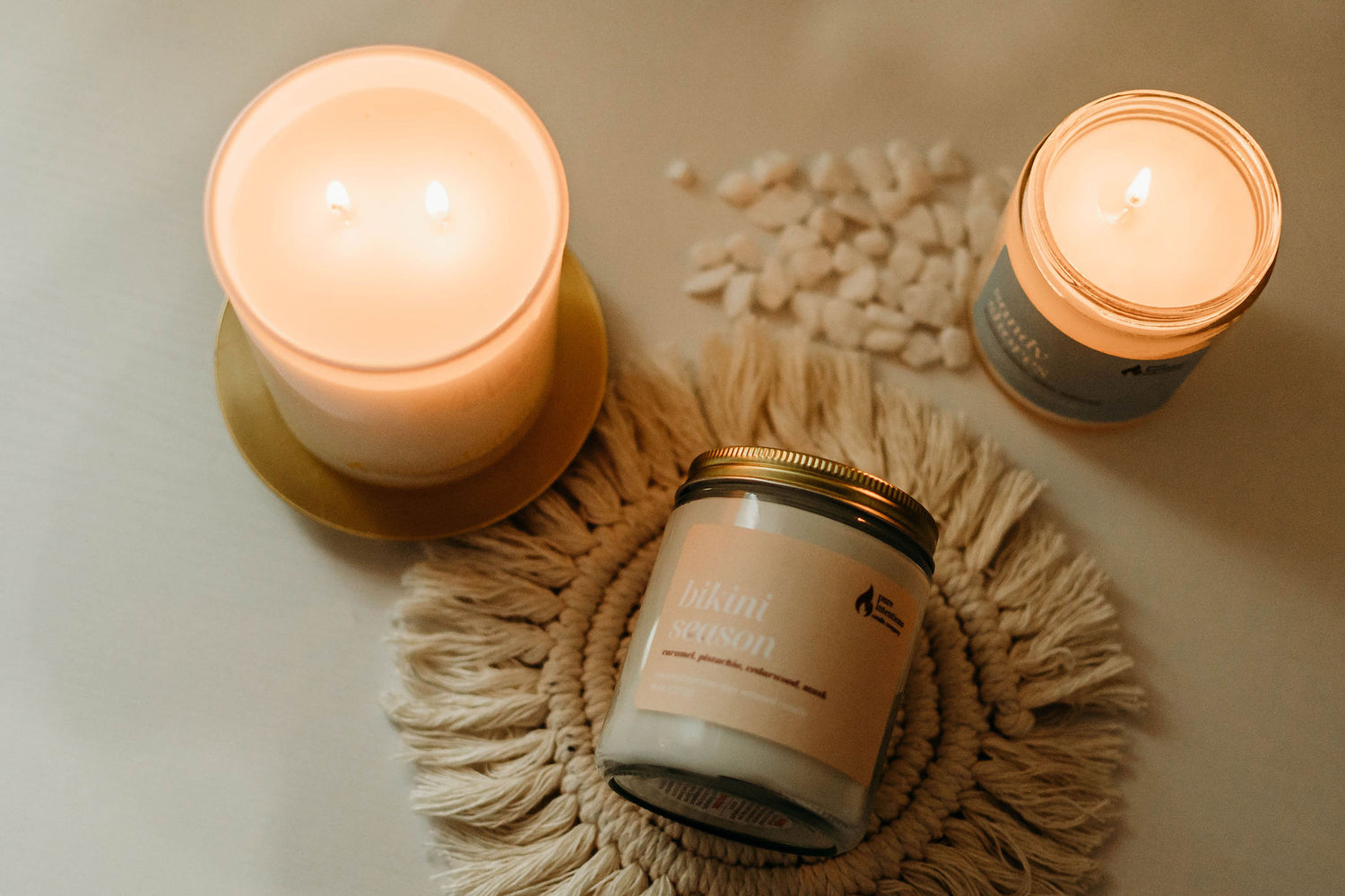 The image shows a cozy scene with three lit candles from Pure Intentions Candle Company. A hand is seen lighting one of the candles with a matchstick. The candles are placed on a light-colored surface with decorative elements, including white stones and a woven mat. The central candle is in a glass jar on a gold-colored stand, creating a warm and inviting ambiance. Two other candles, one labeled "Bikini Season" and the other "Havana Nights," are also visible, enhancing the aesthetic appeal.