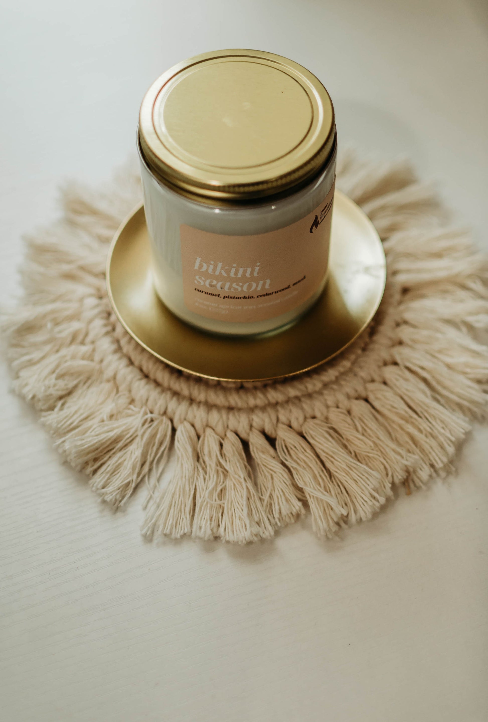 The image displays a single "Bikini Season" candle from Pure Intentions Candle Company. The candle is in a clear glass jar with a gold-colored lid, sitting on a gold plate. The label on the jar is light pink with white text, listing the scent as "Bikini Season" with notes of caramel, pistachio, cedarwood, and musk. The setup includes a decorative, fringed woven mat under the plate, adding a touch of rustic elegance to the scene.