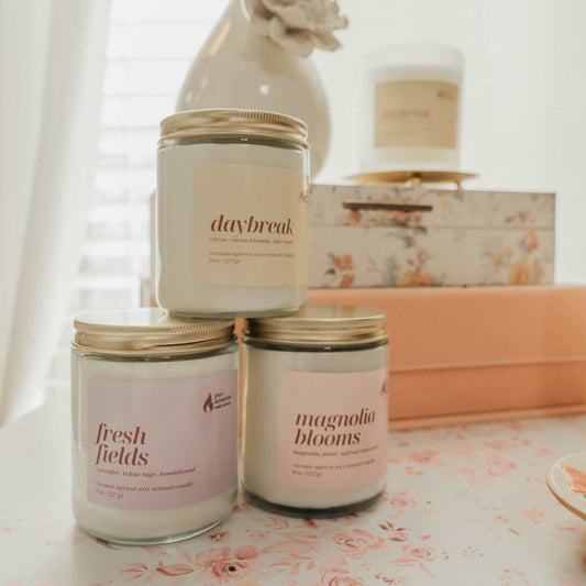 Three candles labeled 'daybreak', 'fresh fields', and 'magnolia blooms' on a floral tablecloth.