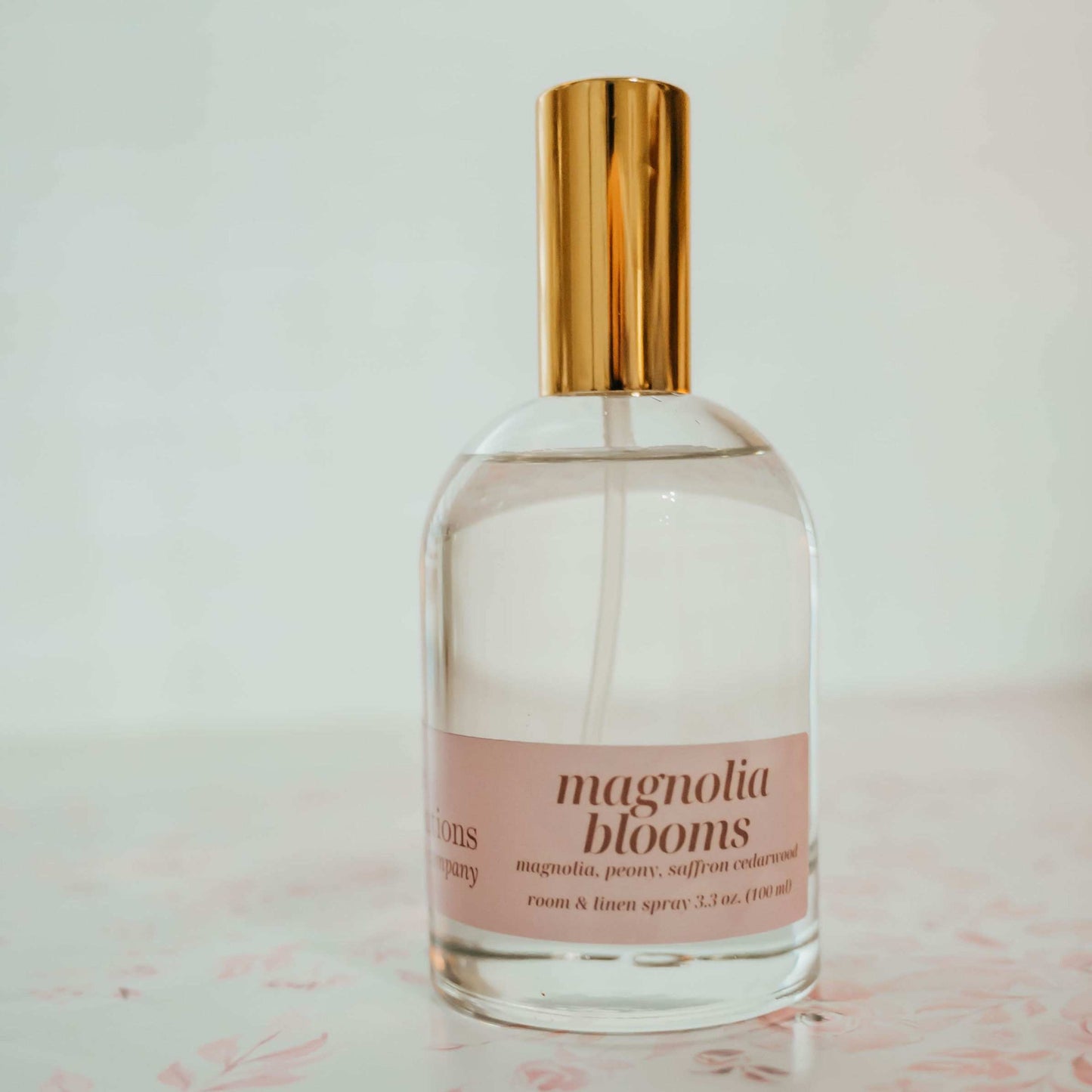 Clear spray bottle labeled ' magnolia blooms' with a gold cap on a light surface.