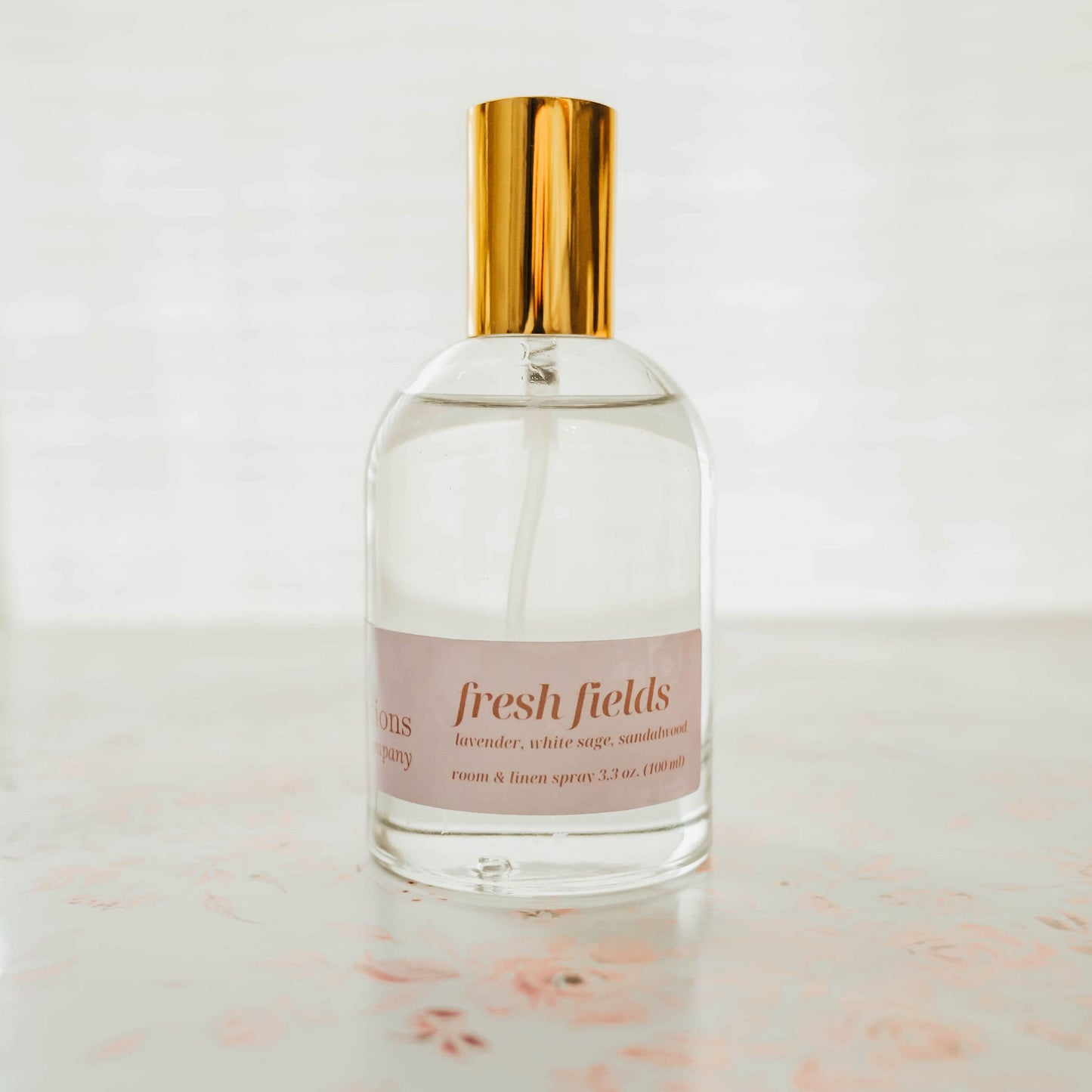 Clear spray bottle with gold cap and pink label on a light surface