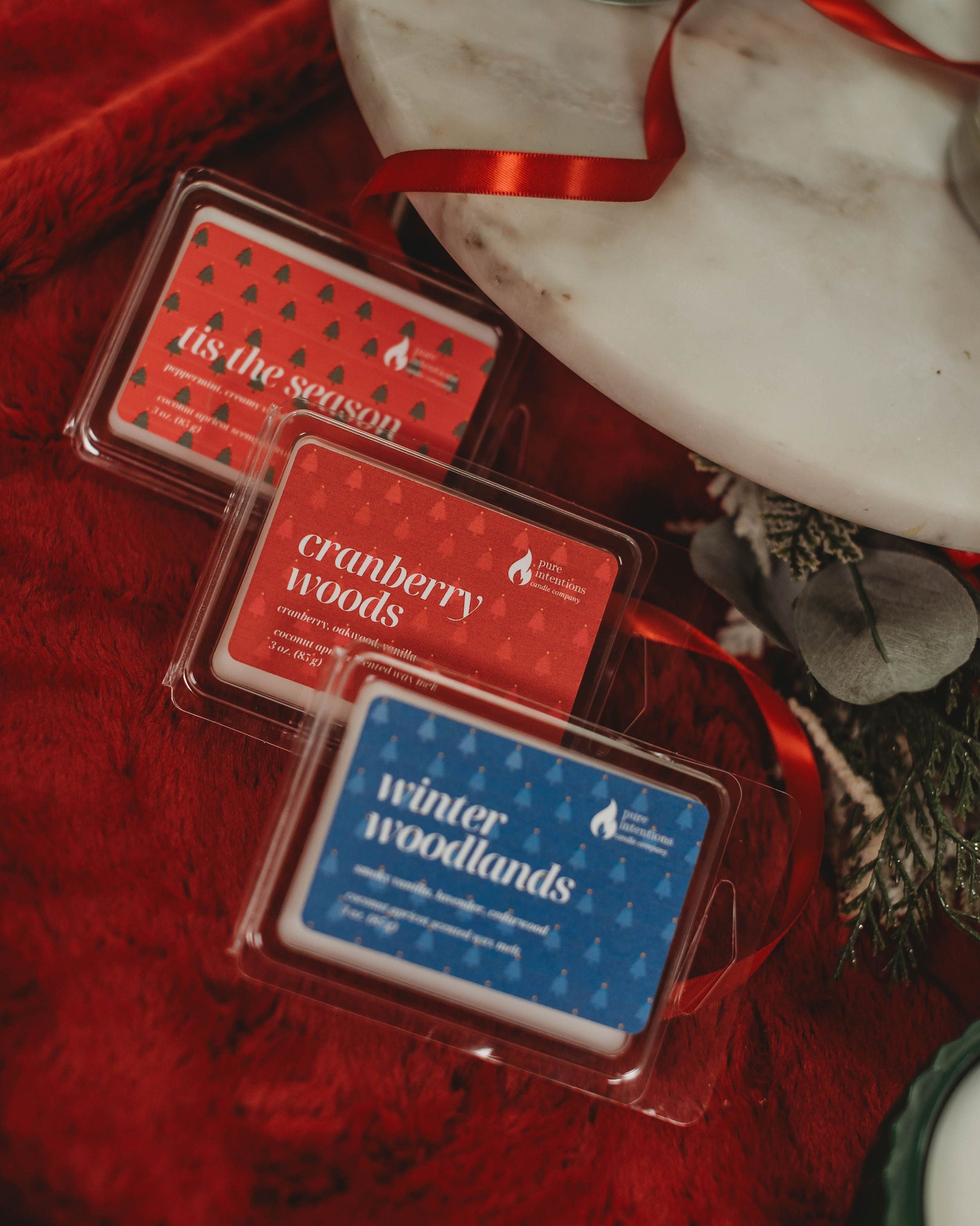 Three wax melts in packaging with 'Cranberry Woods' and 'Winter Woodlands' labels on a red fabric background.