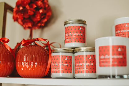 Candles with 'this is the season' labels on a shelf with red decorative items.