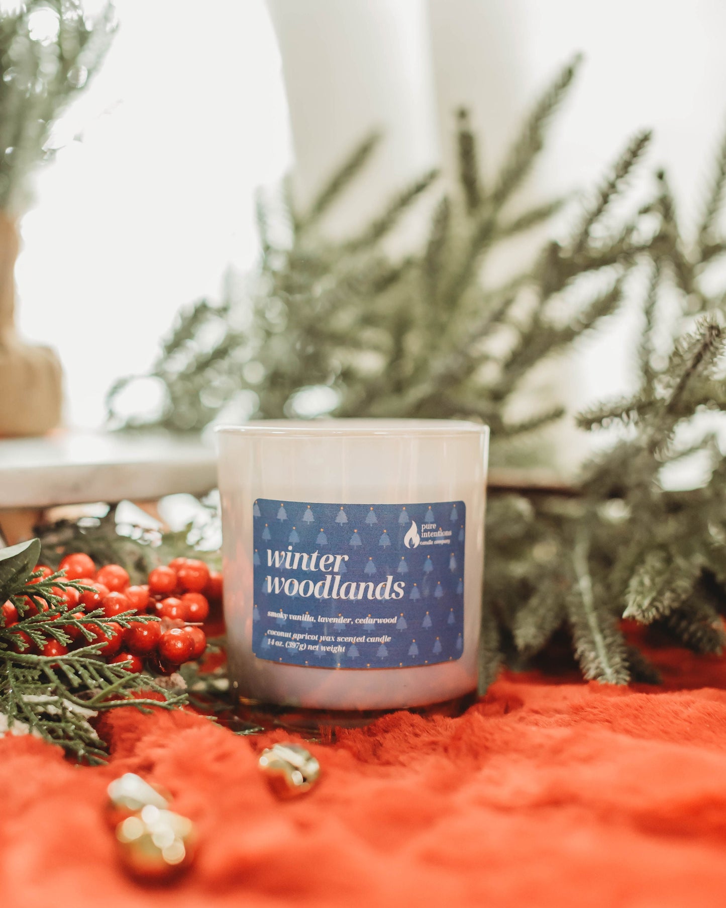 Candle labeled 'winter woodlands' on a red surface with greenery in the background