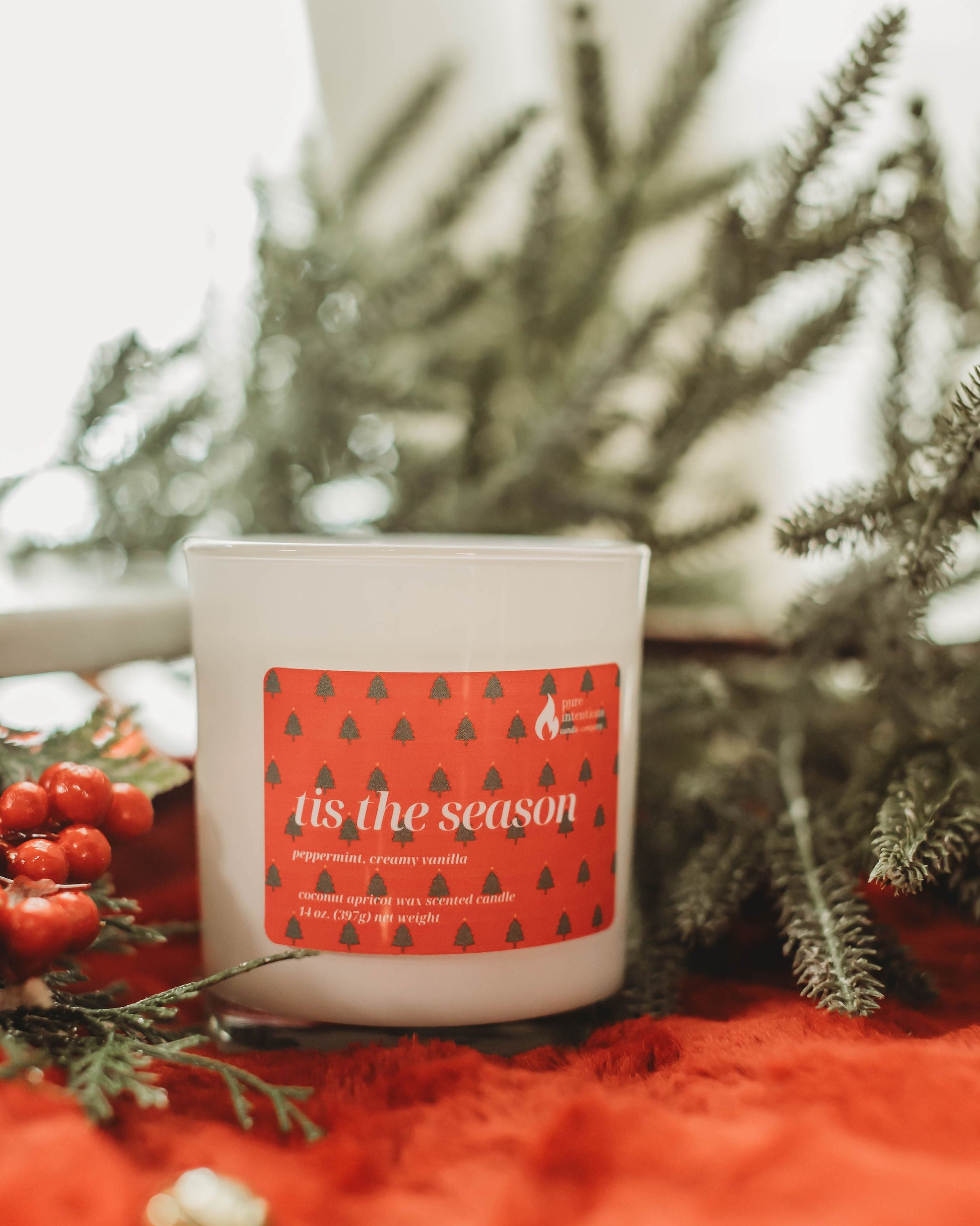 Candle with a 'tis the season' label surrounded by greenery and berries on a red surface.