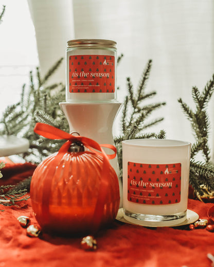 Two candles with 'it's the season' text on a red surface with Christmas decorations.