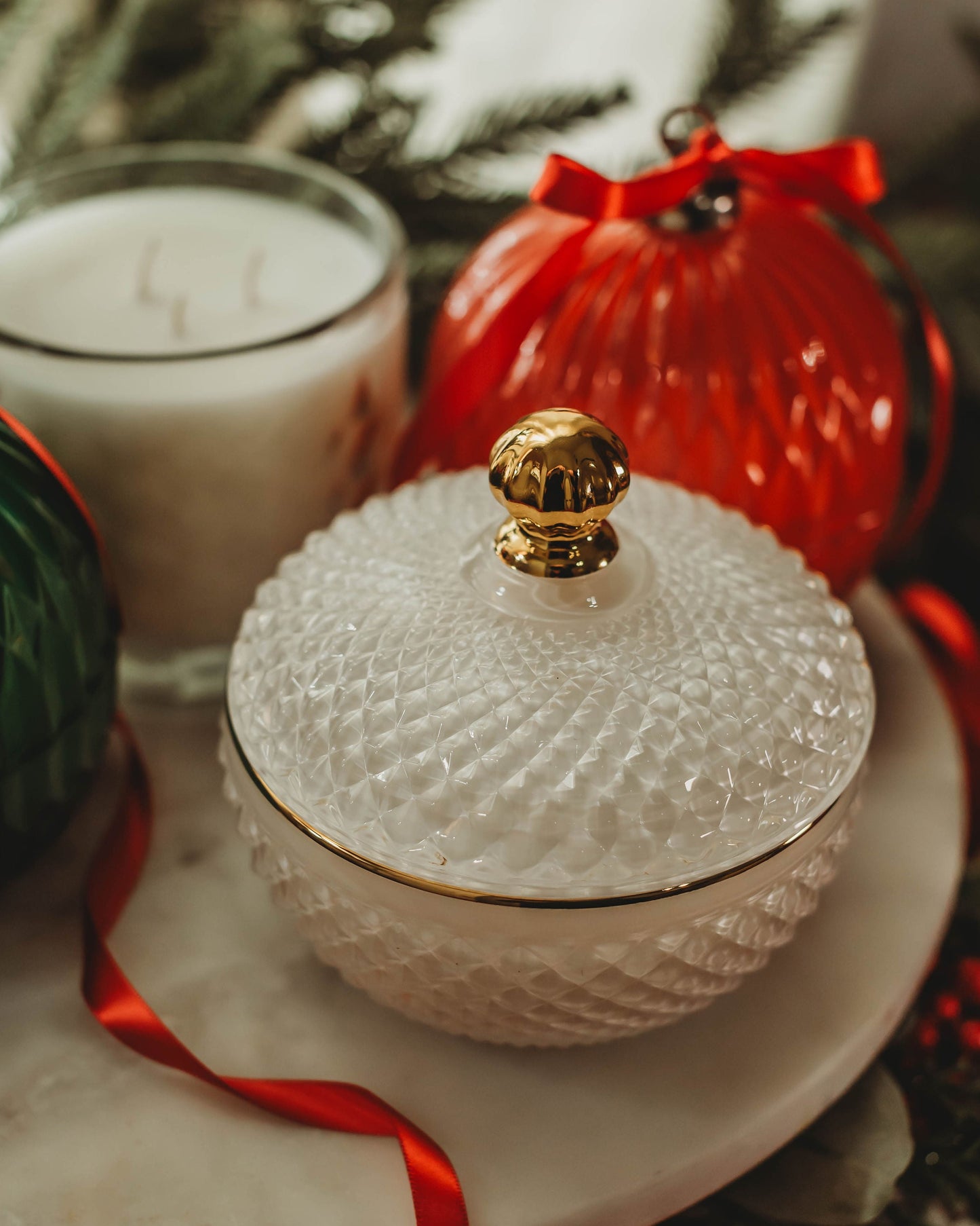 Decorative white textured container with gold lid on a marble surface with red ribbon and blurred greenery in the background.