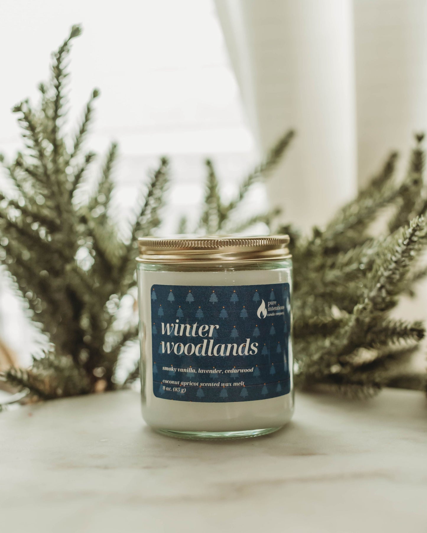 Candle jar labeled 'winter woodlands' on a surface with blurred greenery in the background