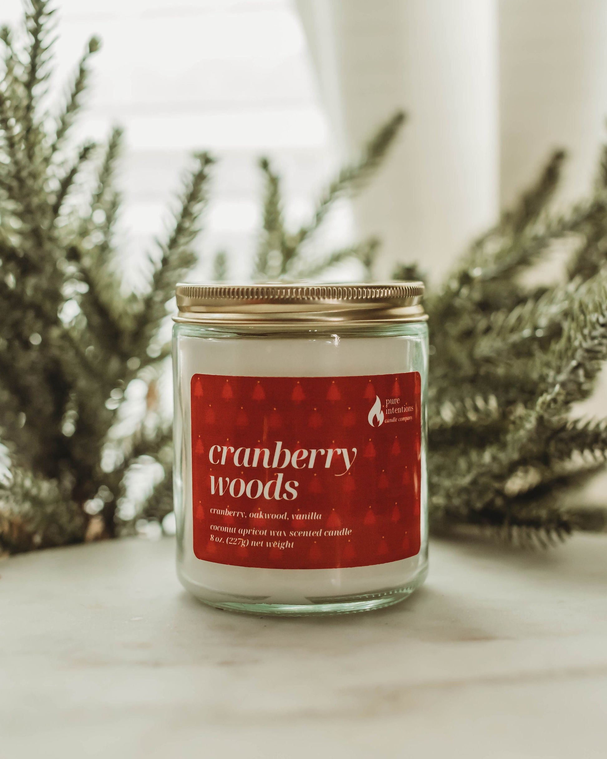 Candle jar labeled 'cranberry woods' with a red label on a surface with greenery in the background.