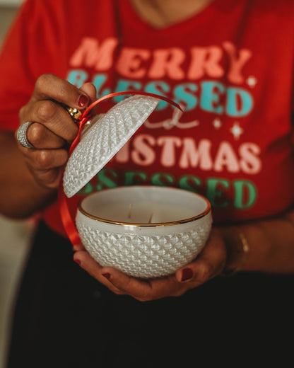 Person holding a white textured candle with a red lid, wearing a red shirt with Christmas text.
