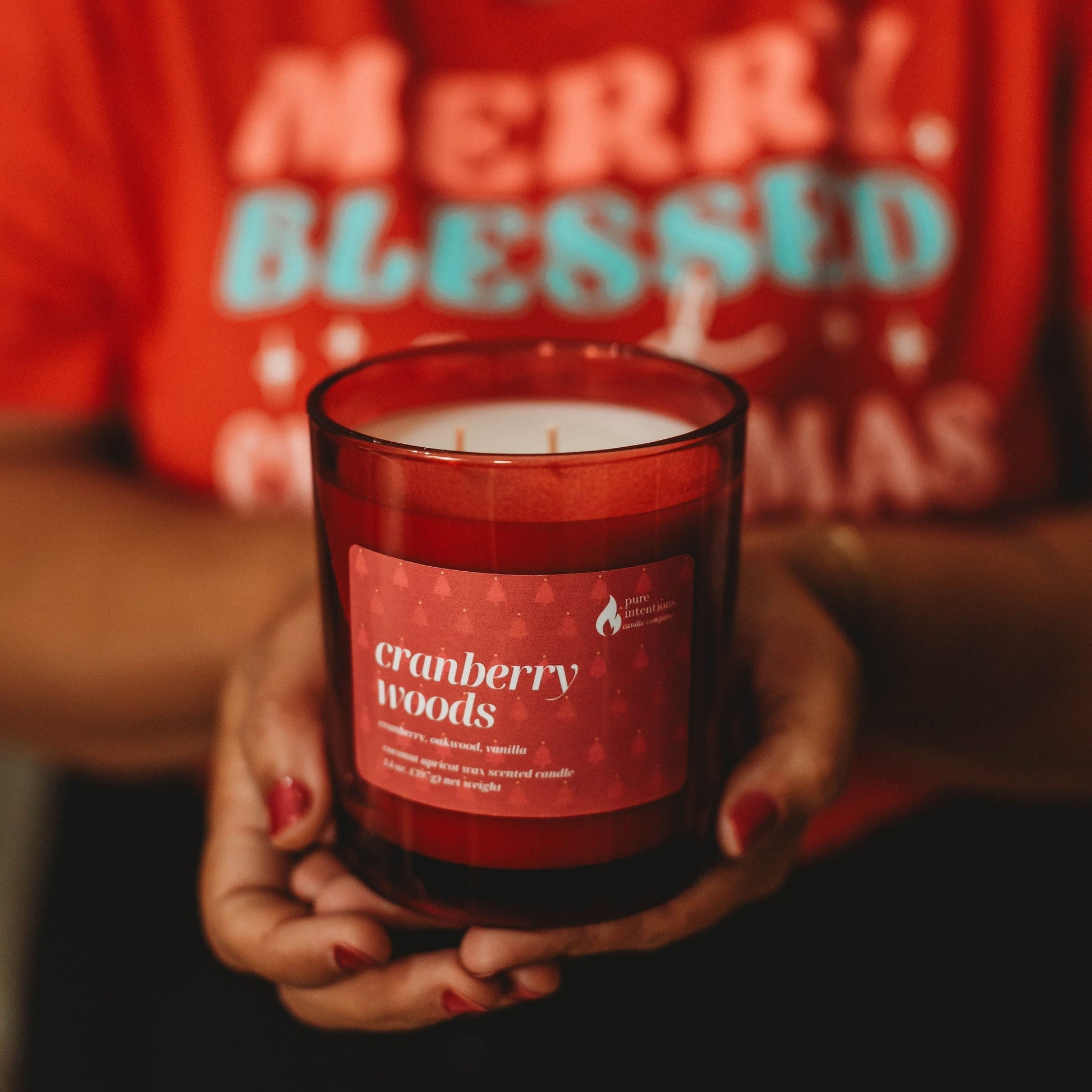 Person holding a 'Cranberry Woods' candle with a red shirt in the background