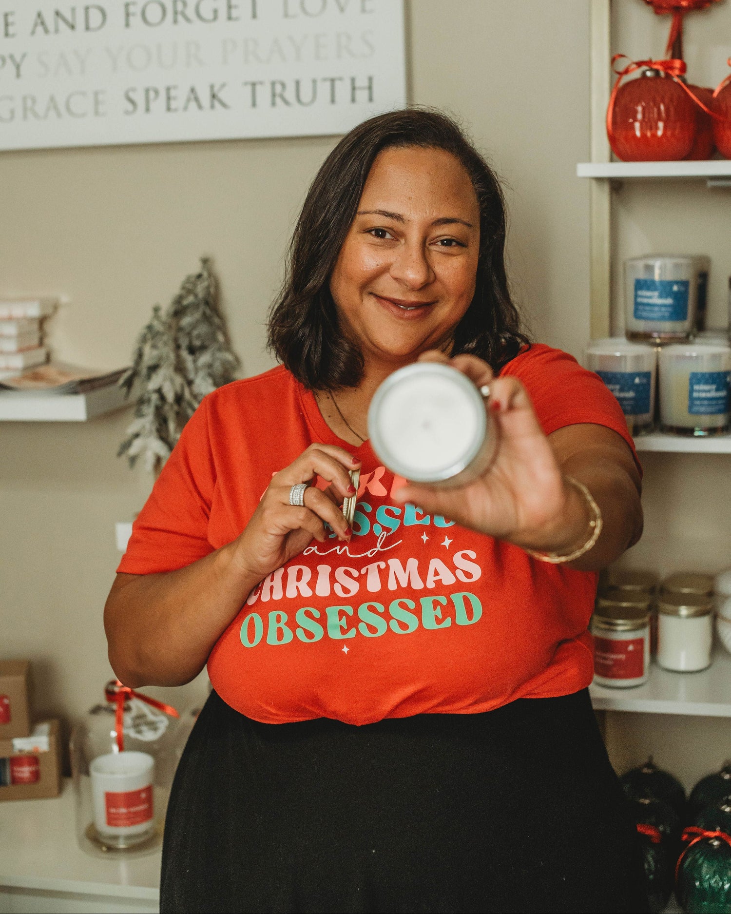 Woman in a red 'Christmas Obsessed' shirt holding a Christmas candle in a front of candles. 