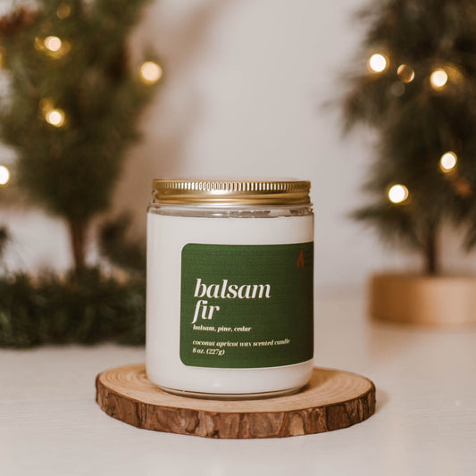 Balsam Fir candle from Pure Intentions Candle Company in an 8 oz. jar, featuring a green label with the scent notes of balsam, pine, and cedar. The candle is placed on a wooden coaster, surrounded by a cozy holiday background with blurred pine trees and warm string lights