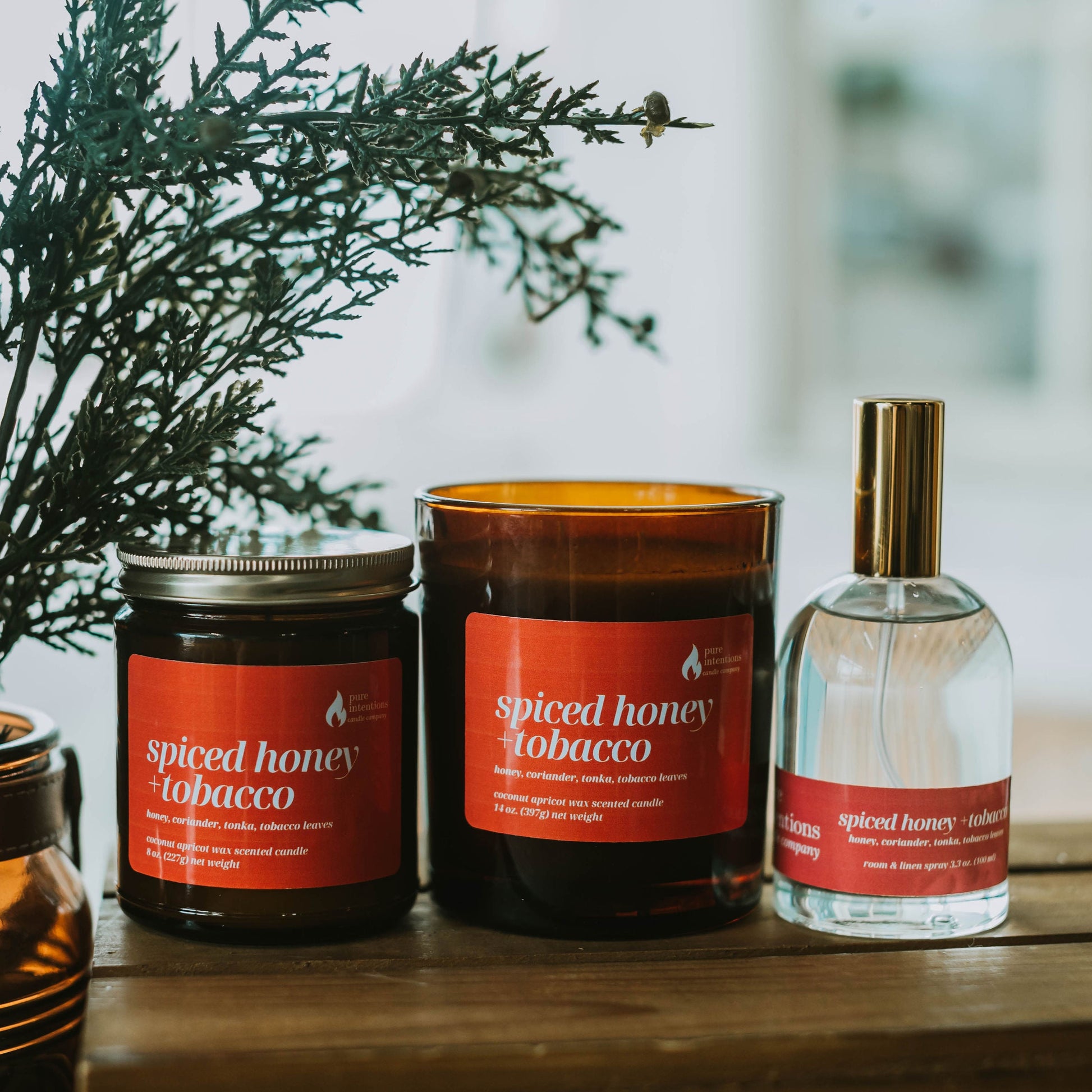 Spiced honey tobacco candles and a perfume bottle on a wooden surface with greenery.
