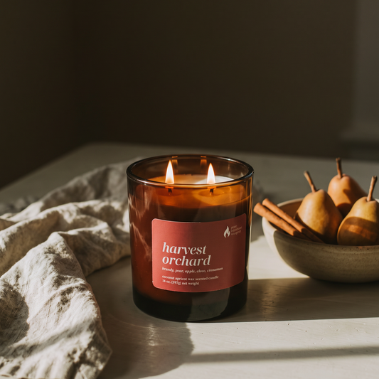 A lit amber glass candle with a terracotta label reading 'harvest orchid' sits on a tabletop beside a crumpled cloth and a bowl of pears and cinnamon sticks in warm natural light.