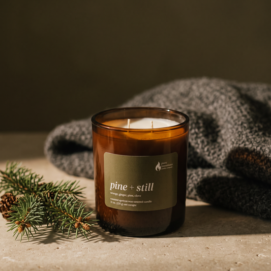 An amber glass jar candle labeled 'pine + still' sits on a stone surface with a folded gray knit blanket behind it and pine branches in the foreground, creating a cozy styled product scene.